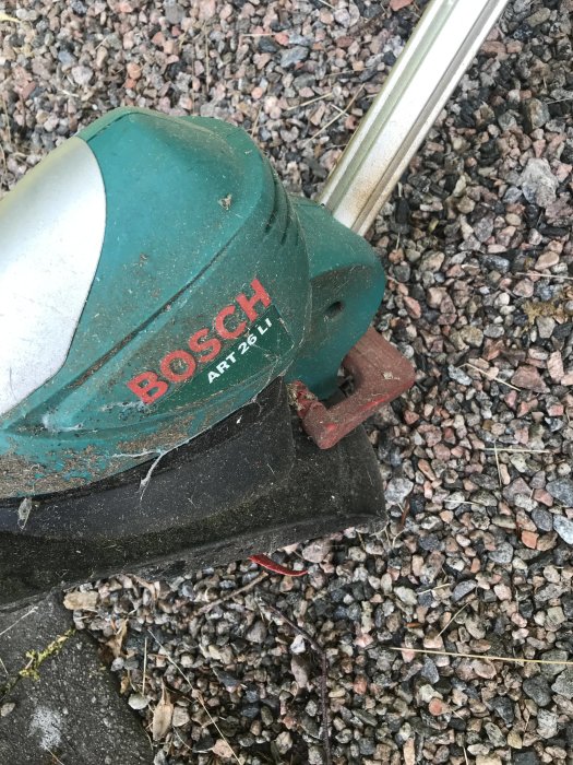 Delvis täckt av damm visar bilden en Bosch ART 26 Li batteridriven trimmer liggande på grus.