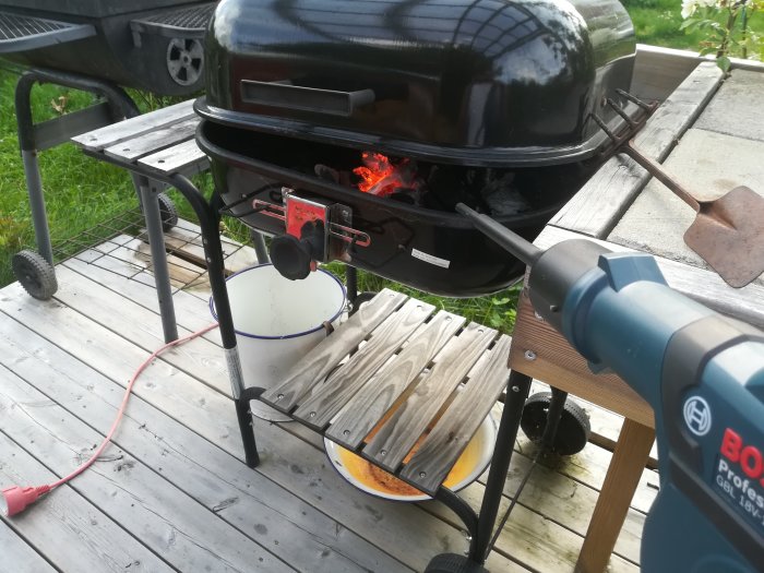 En grill med öppet lock och synliga glödande kol, bredvid en byggfläkt och spackelspadar på en trädäck.