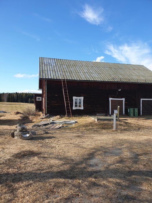 En gammal timrad ladugård med metalltak och en stege lutad mot fasaden, omgiven av en gräsmatta.