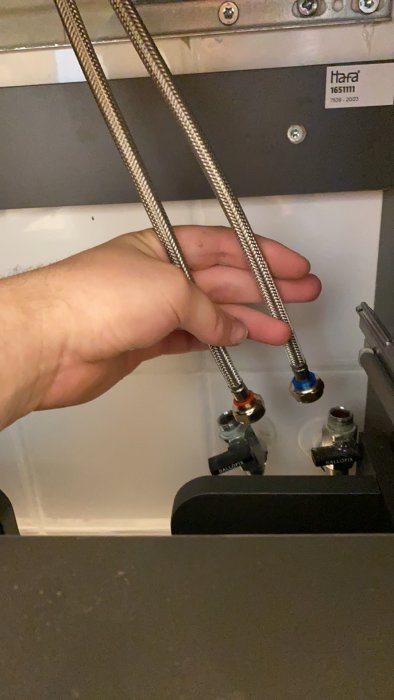 Hand håller två för korta anslutningsslangar som inte når vattenkranens anslutning under ett handfat.