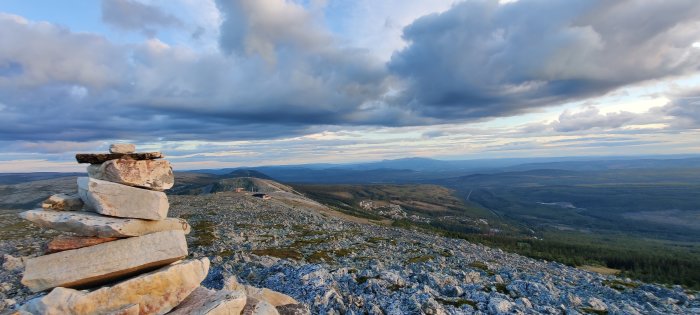 Stenig fjälltopp med stencirklar och utsikt över ett skogbeklätt landskap vid skymning.