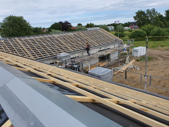 Hus under konstruktion med halvt lagt tak och installation av solcellsskenor, omringat av byggnadsställningar.