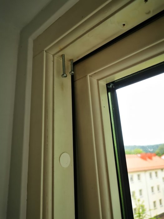 Skruv delvis inskruvad i fönsterramens övre del med öppet fönster och utsikt över byggnader i bakgrunden.