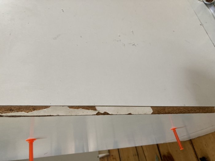 Skadad kant på en skiva där plastlist ska appliceras, med orange spik som stöd underifrån.