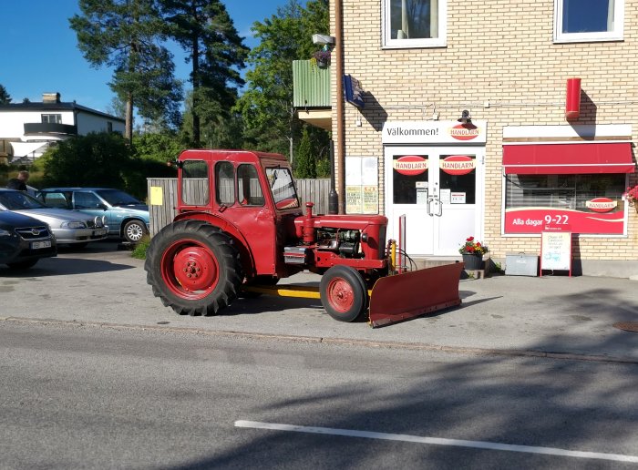 Röd traktor med snöplog parkerad utanför en livsmedelsbutik under sommaren.