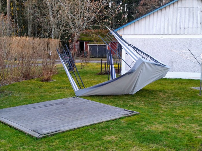 Skadat partytält med böjda stänger och löst tyg som blåst ner på en gräsmatta vid en träveranda.