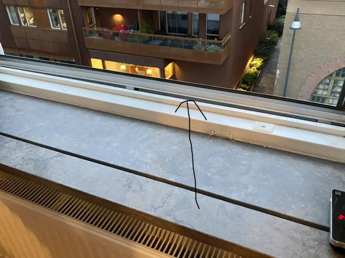 Böjd fönsterskena i vit färg som sitter fast på en fönsterkarm med utsikt mot bostadskomplex.