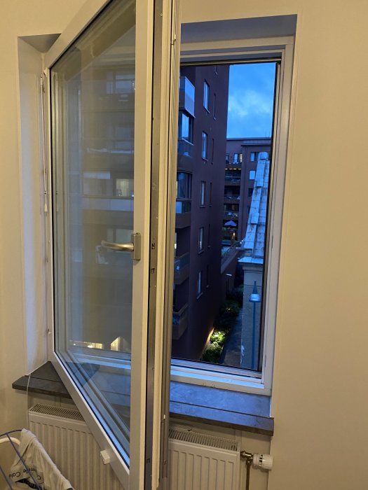 Öppet fönster med en böjd skena synlig, i en lägenhetsmiljö vid skymningen.