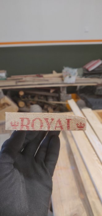 Hand i svart handske håller en träskurkloss med hyvlad kant och texten "ROYAL" stämplat i rött.