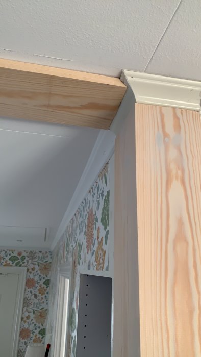 Hörndetalj av ett rum med taklister och en obehandlad träpelare vid en tapetserad vägg.