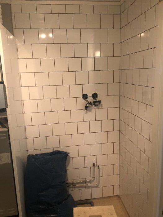 Renoverad vägg i tvättstuga med halvkaklade vita plattor och synliga orörda rör samt pågående arbete.