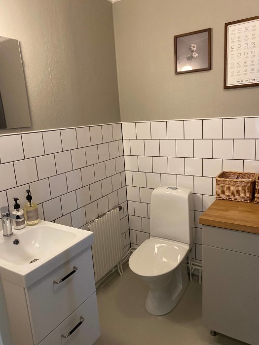 Renoverad gästtoalett med halvkaklade väggar i vitt och gråmålade ovankanter, vit toalettstol och handfat.