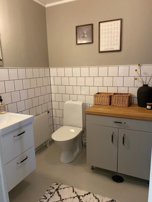 Renoverad gästtoalett med halvkaklade väggar, vit toalett, grått skåp och matchande matta.
