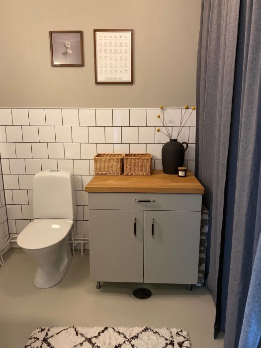 Renoverad gästtoalett med halvkaklade väggar, grålackerat skåp och målad våtrumsmatta.