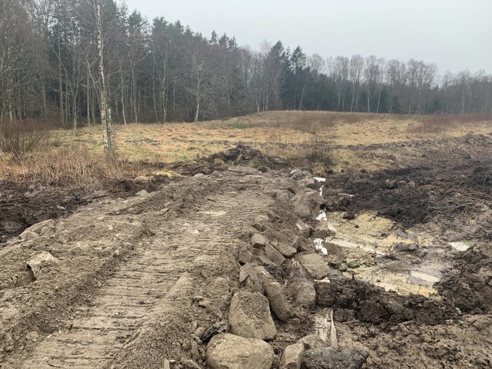 Ett pågående vägbygge med en nygrävd grusväg och diken där rör har lagts, i ett öppet landskap med trädstånd i bakgrunden.