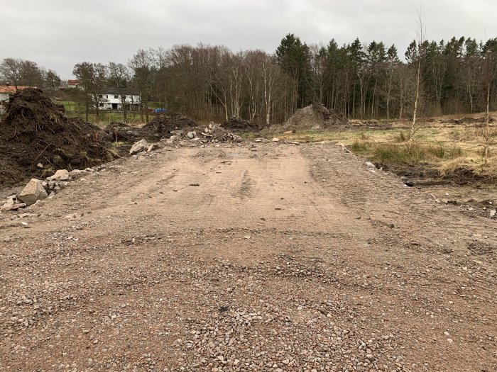 Vägbygge på tomt med grus och högar av jord där framtida garage/verkstad planeras, markerat med pinnar.