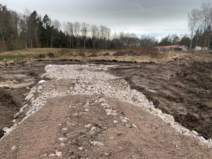 Grusad vägbyggnad mot ett framtida garage med markerade pinnar och rör på en tom byggtomt.