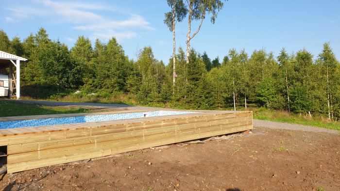 Halvt nedgrävd hemmabyggd pool omgiven av trädäck med skog i bakgrunden på en klar dag.