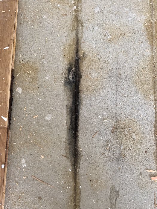 Uppbrutet parkettgolv som visar en underliggande betong med en långsträckt svart fläck och spånrester.