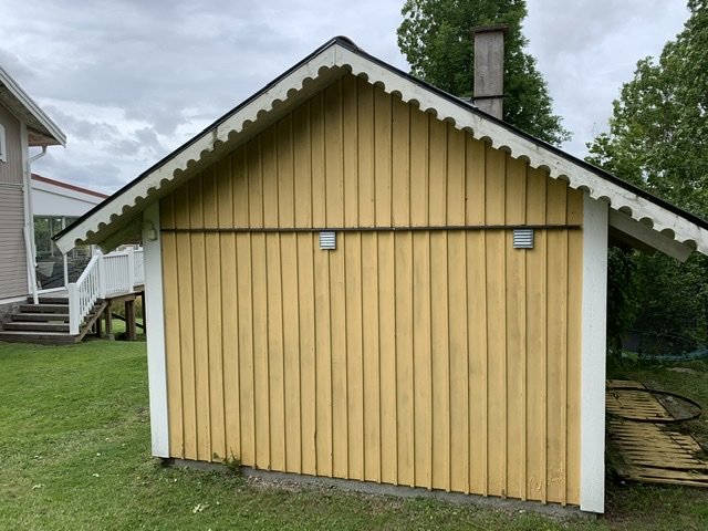 En gulmålad lillstuga med liggande panel och en vitmålad hörnlist, två ventilationsgaller och grönt gräs framför.