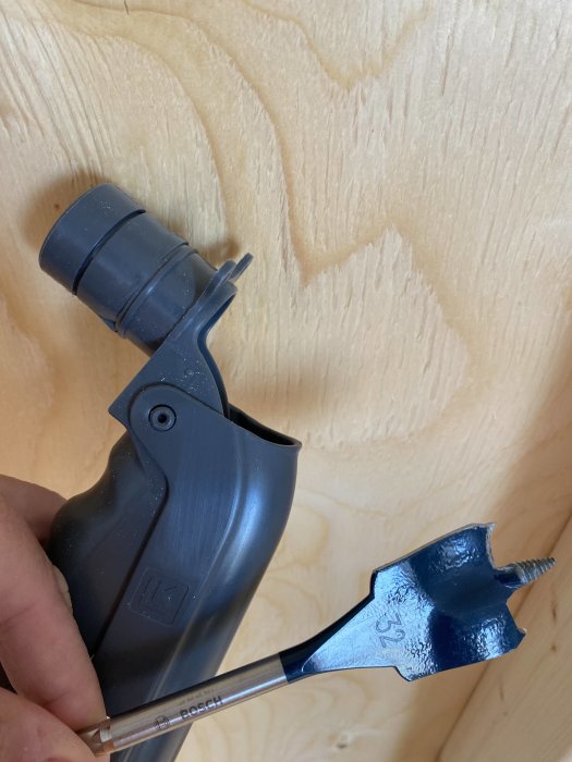 En hand håller en LK bockfixtur nära en plywoodvägg med en 32 mm hålsåg fäst vid en borrmaskin.