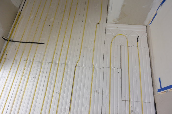 Vit skumisolering ordnad på betonggolv med gul elkabel liggande ovanpå, beredd för golvvärme installation.