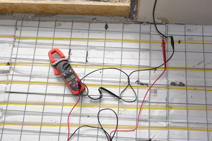 Installation av golvvärmesystem med en gul elkabel som testas med en digital multimeter på en isolerad betongyta.