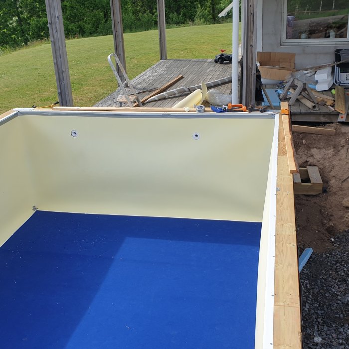 Inbyggd pool under konstruktion med plywoodväggar, blå matta i botten och hål för vattenanslutningar.
