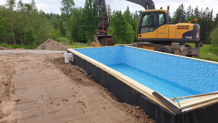 Grävmaskin fyller jord vid pool som är under konstruktion med blå klinker och synligt avloppsrör.