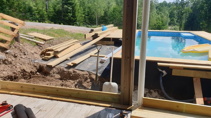 Byggprocess av trädäck nära pool med grävd grop för vattenpump och trall som läggs.