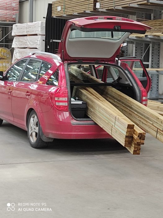 Röd bil överlastad med långa träplankor som sticker ut från öppen baklucka i ett varuhus.
