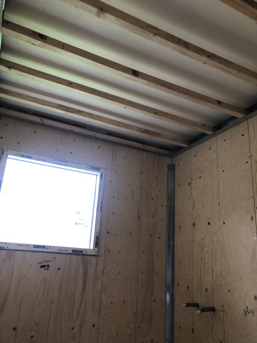 Interiör av ett rum under renovering med plywoodväggar och sänkt tak med synliga träreglar.