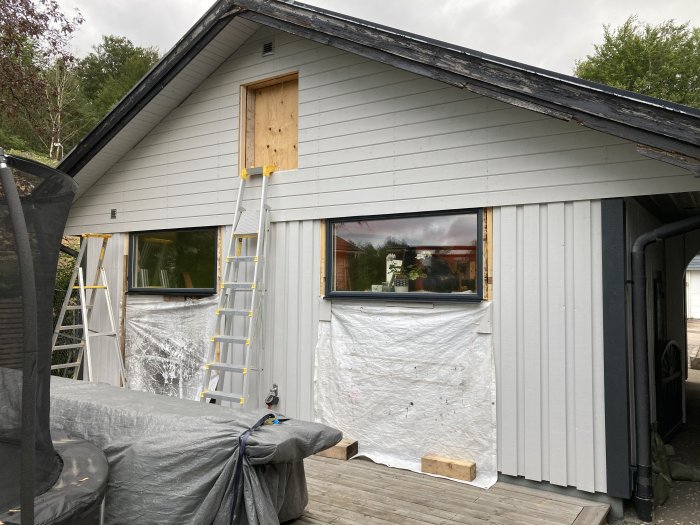 Husfasad med nyinstallerad panel och en stege lutad mot väggen, skyddsplast på delar av byggnaden.