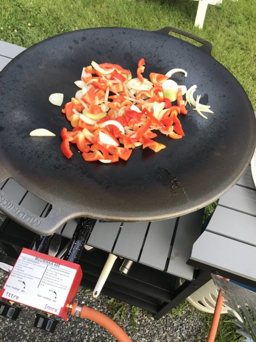 Stekhäll med paprika och lök under tillagning står på en ny gasolgrill utomhus, med en broschyr och köksredskap synliga.