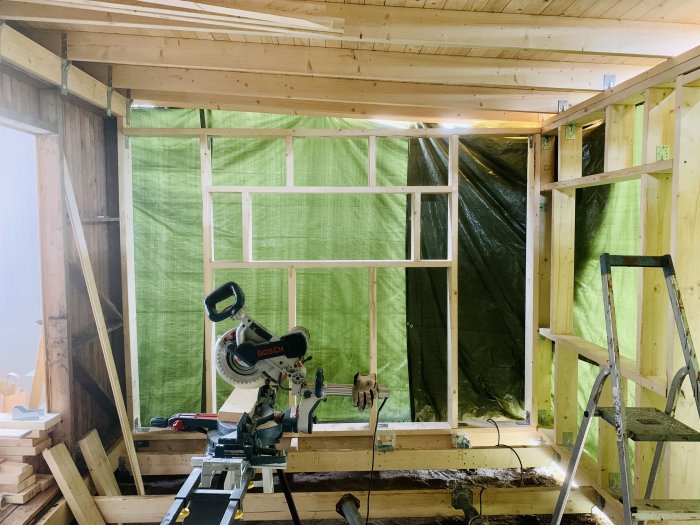 En pågående renovering inne i ett trähus med synliga reglar, överdragna med vindskydd och byggverktyg.