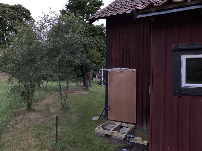 En hemmagjord utomhusdusch med draperi och trall vid en röd stuga, med grönskande trädgård och dasset i förgrunden.