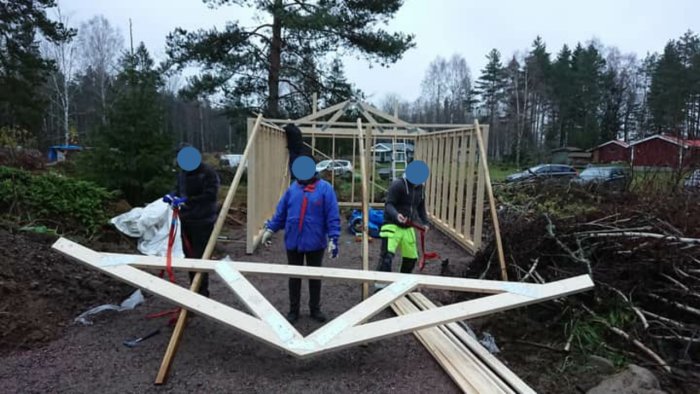 Byggande av en trästomme för hus med en W-formad takstol framför och personer som arbetar.