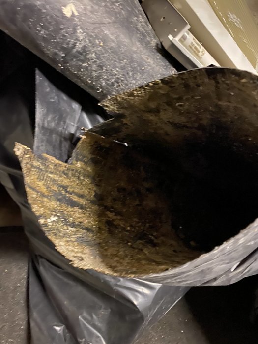 Närbild av en skadad del av en svart plastdurk med tydliga skador och repor, placerad i ett mörkt garage.