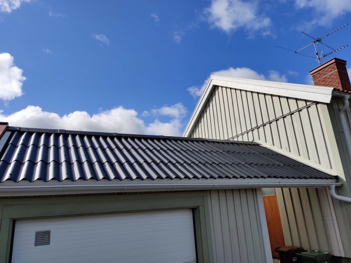 Nytt grått fibercementtak på garage, bredvid beige husvägg med blå detalj, under blå himmel.