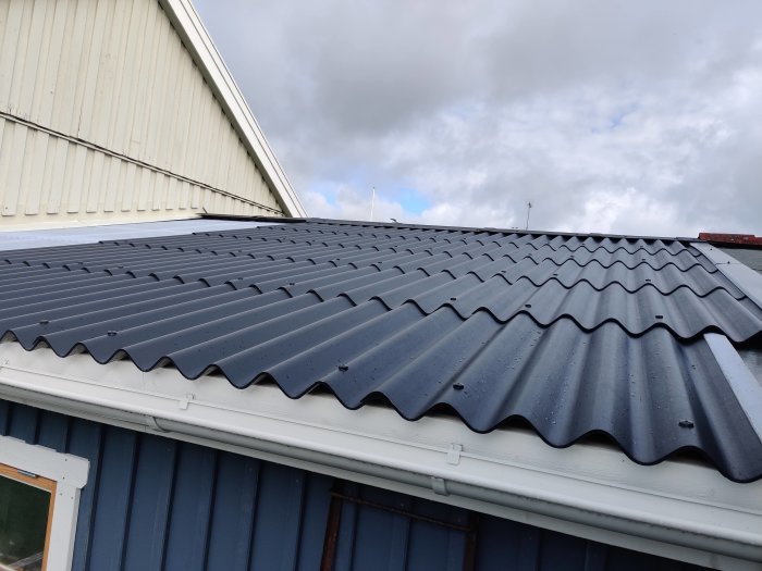 Nytt grått vågigt fibercementtak monterat på garage, med plåtdetaljer och befintlig byggnadsstruktur i bakgrunden.