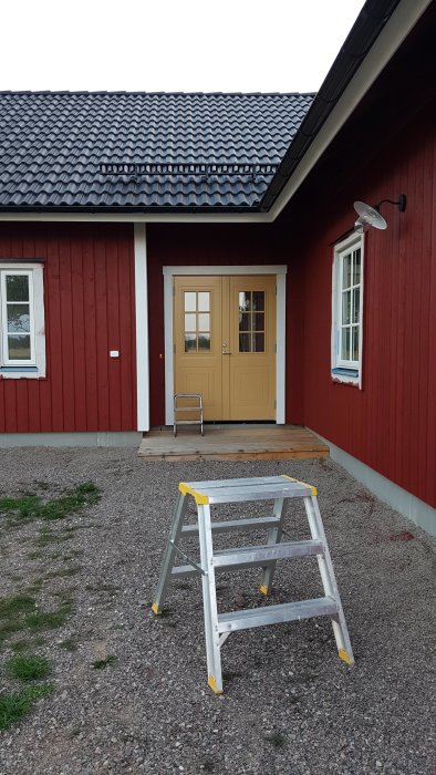 En trappa framför ingången till ett rött hus med vita knutar och en gul dörr.