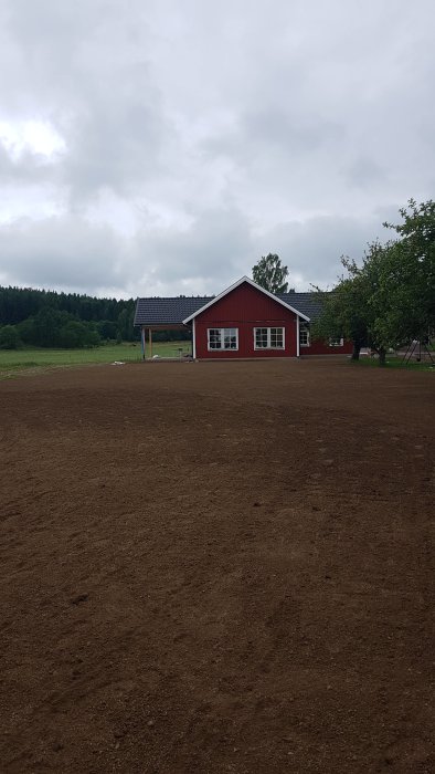 Rött enfamiljshus med vita fönsterkarmar omgivet av nyplöjd mark under en molnig himmel.