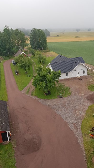 Vy över en lantlig fastighet med ett enfamiljshus omgivet av gröna träd och en grusdrivväg.