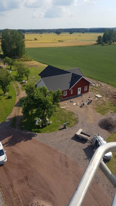 Vy över landskap med rött hus och svart tak, grusväg och leksaker på gården, tagen från hög vinkel.