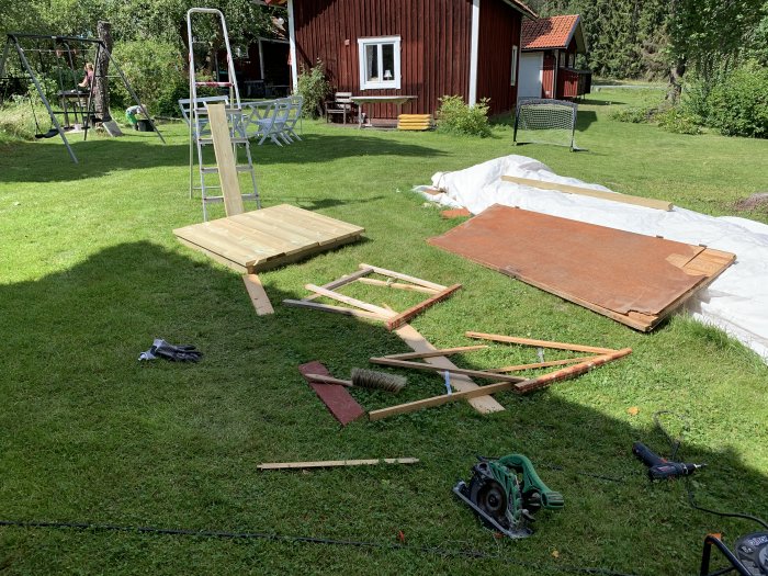 Sammansatt träarbetsbord som kollapsat på gräsmatta framför rött hus, med verktyg och lösa trädelar omkring.