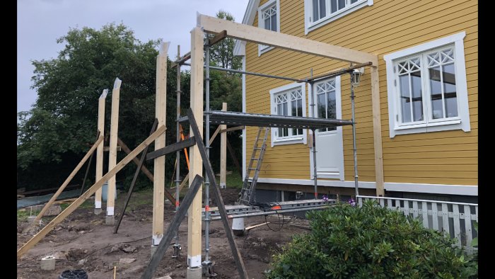 Nyinstallerad träbalk på bygge framför gult hus med stödställningar och byggmaterial runtomkring.