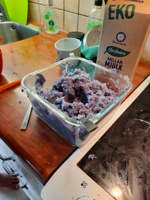 Mosad blå potatis i en genomskinlig glasskål på ett köksbord med en mjölkförpackning bredvid.