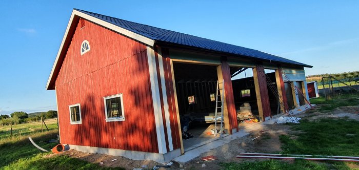 Röd träfasad på byggnad under konstruktion, plåttak, lecablock och öppen carport, byggmaterial synligt.