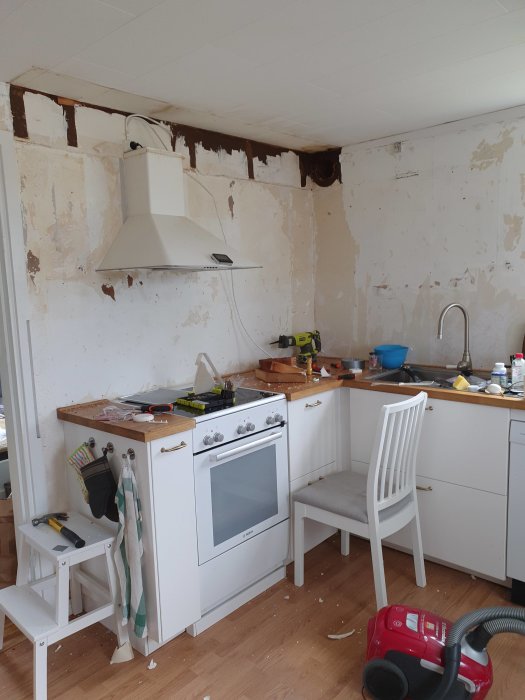Ett kök under renovering med osammanhängande vita skåp, en oavslutad vägg och en fristående utsugsfläkt.