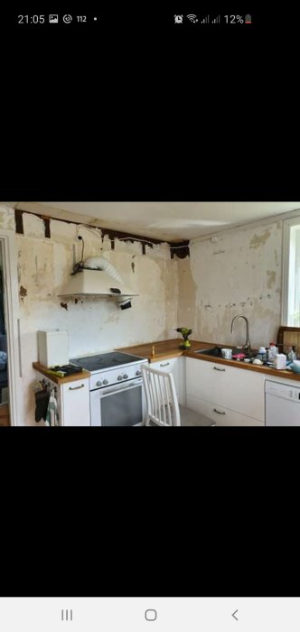 Kök under renovering med avflagad väggfärg och en fläkt som saknar utblås, kopplad till ett synligt rör som leder mot taket.